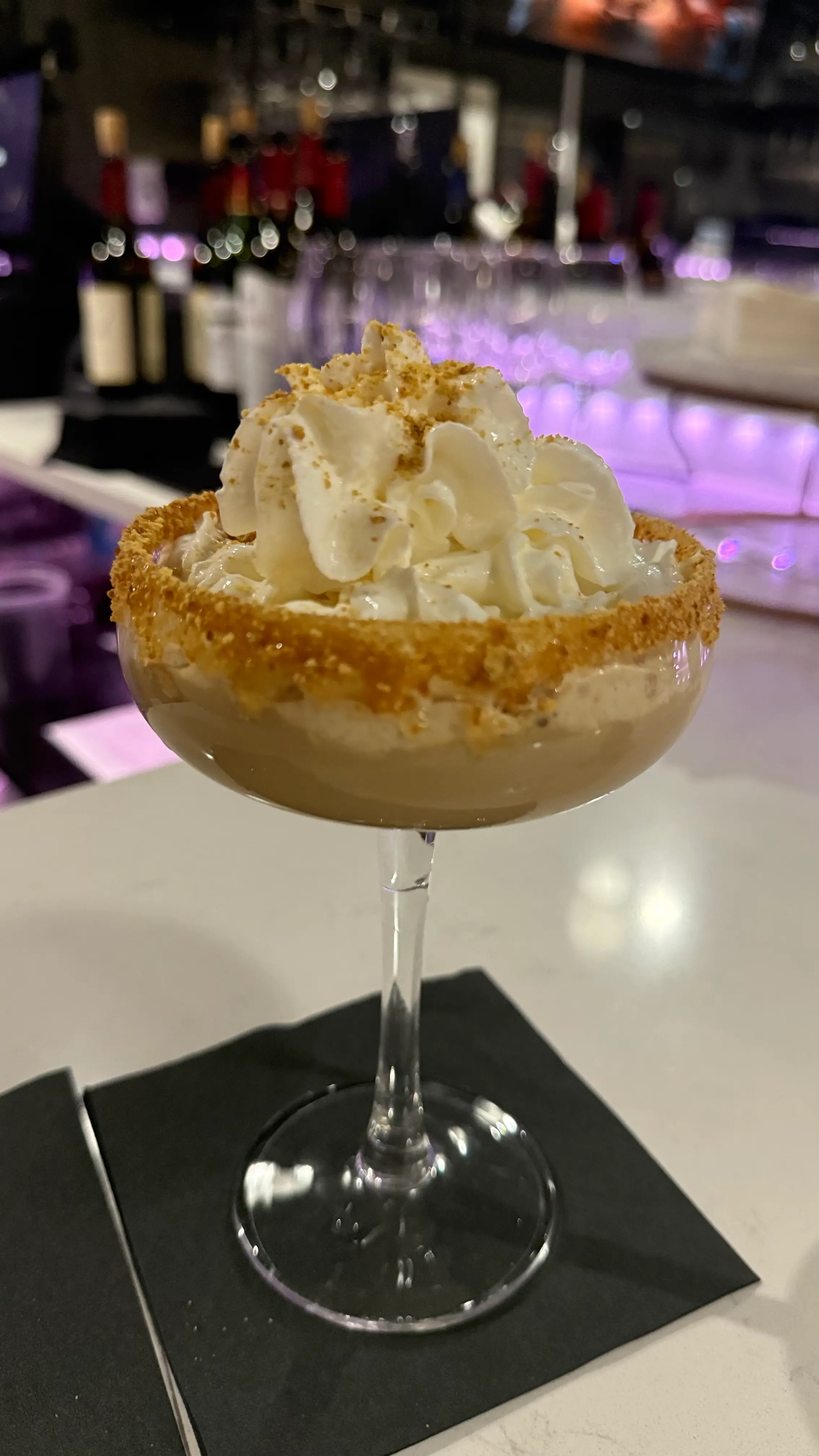 Cocktail glass with creamy drink topped with whipped cream and crushed biscuit rim, placed on a black napkin at a bar.