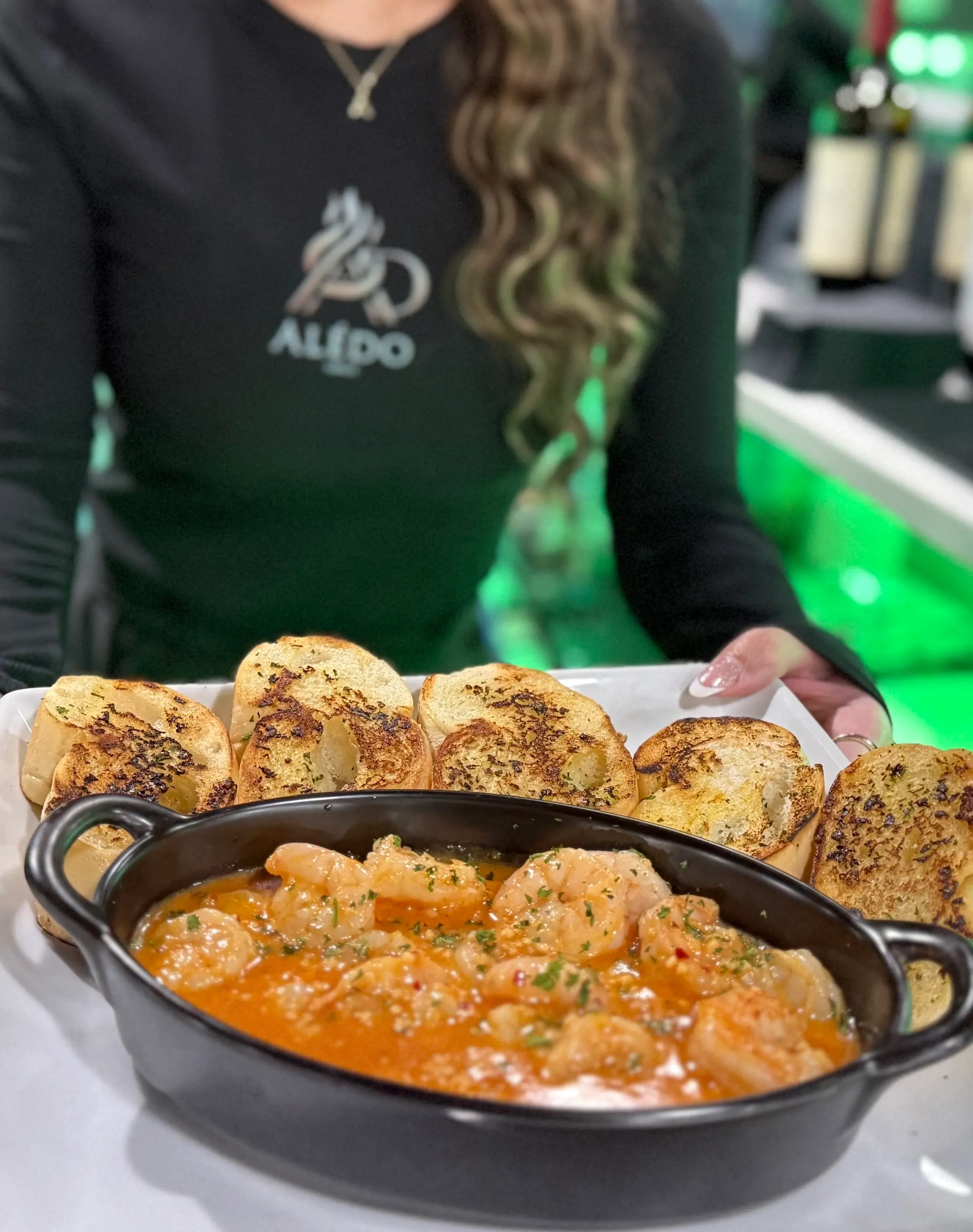 Black oval dish of shrimp cooked in red sauce, served with toasted bread slices on a white tray held by a person in a black shirt.