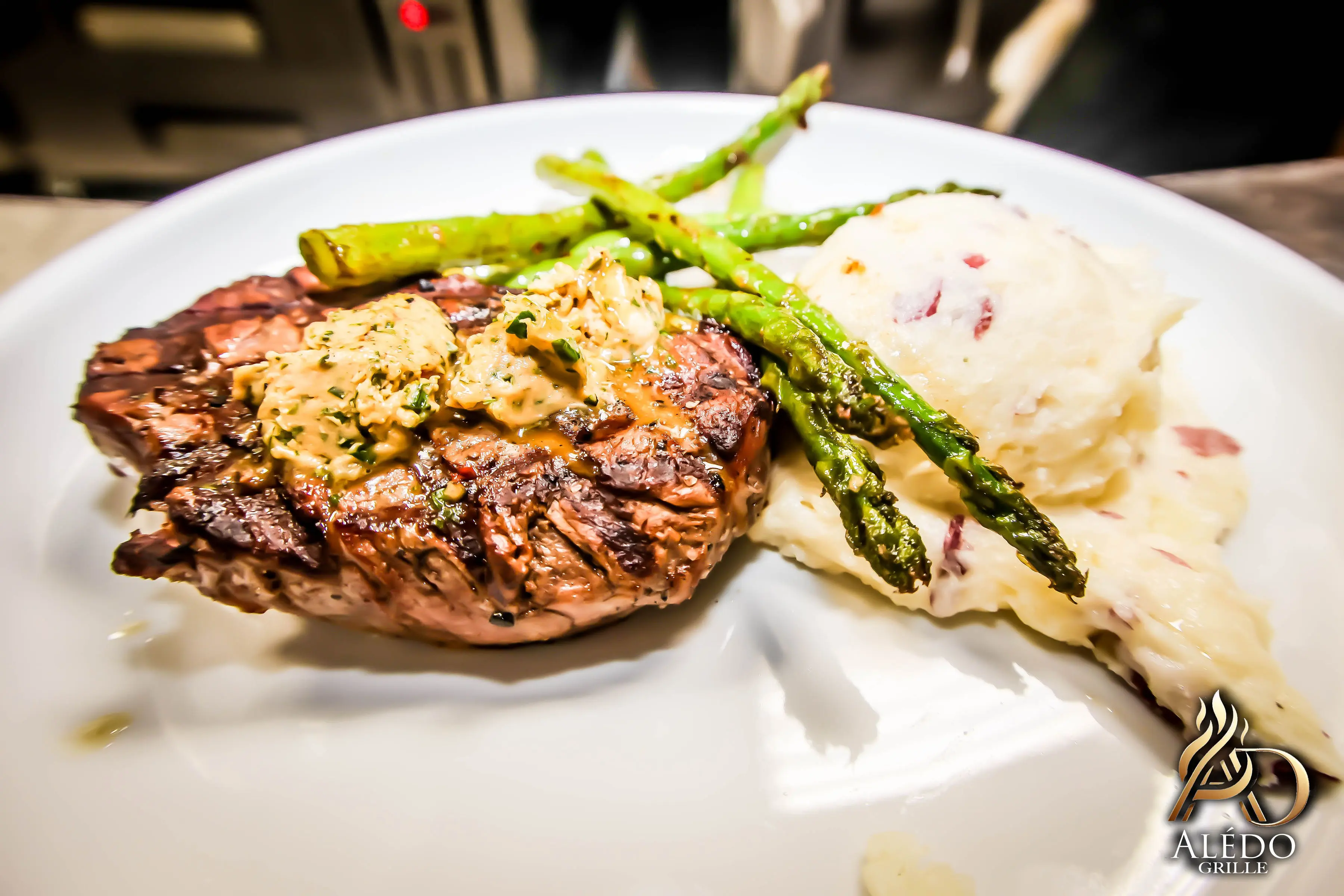 Grilled steak topped with herb butter, served with roasted asparagus and creamy mashed potatoes on a white plate.