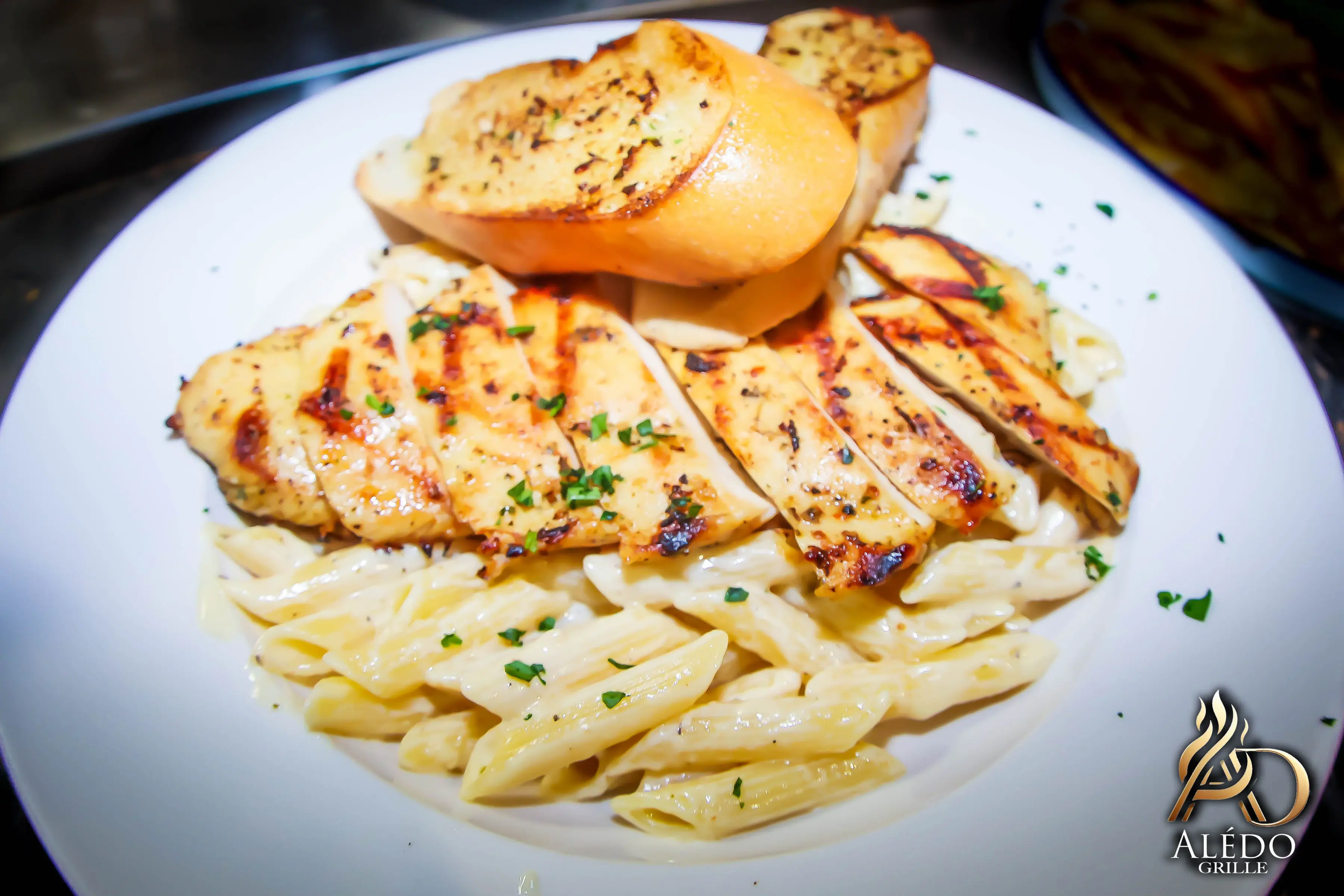 Grilled chicken breast slices served on creamy penne pasta with two pieces of garlic bread, garnished with chopped parsley.