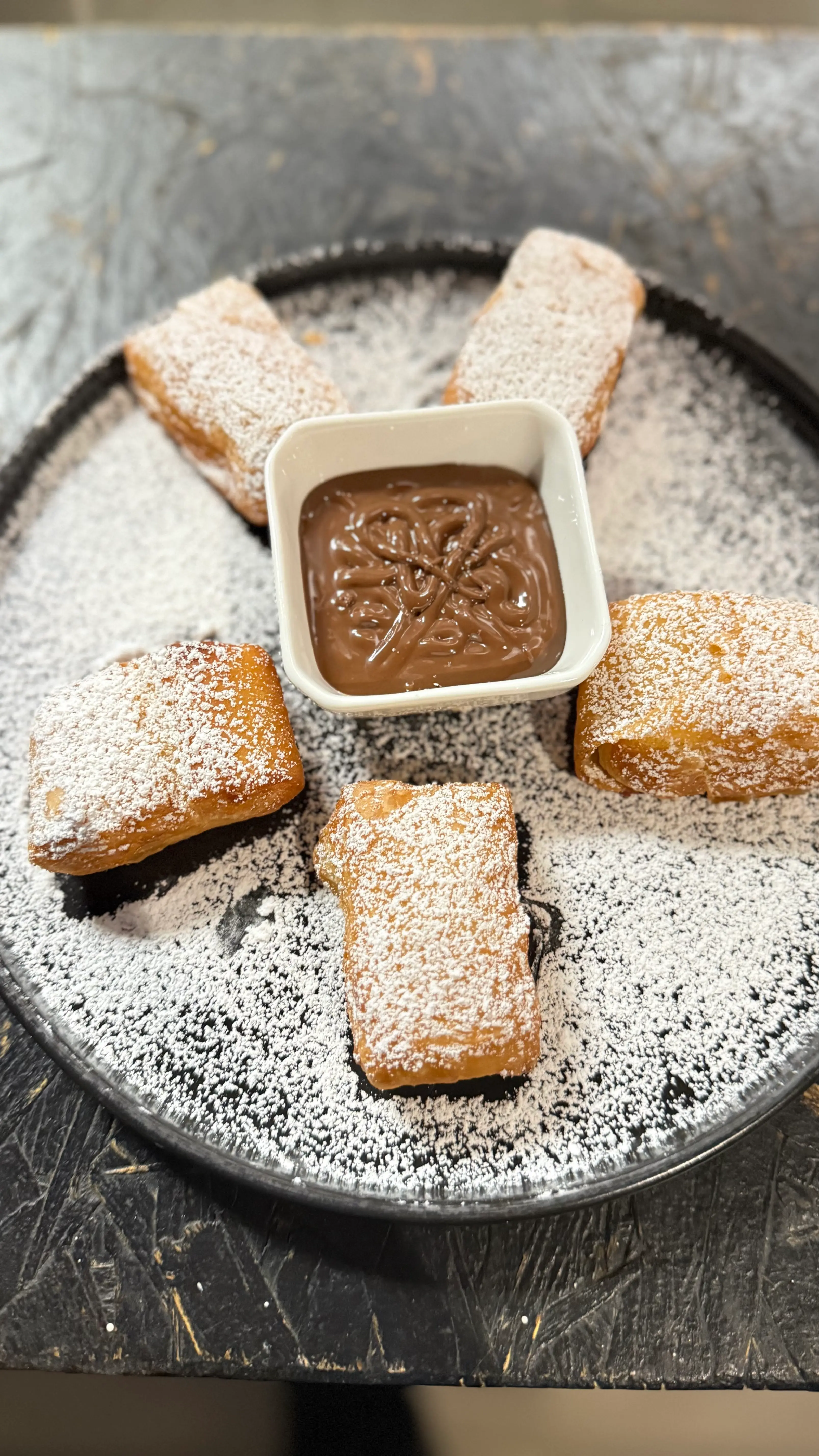 Plate with five powdered sugar-dusted fried pastries surrounding a square bowl of chocolate sauce.