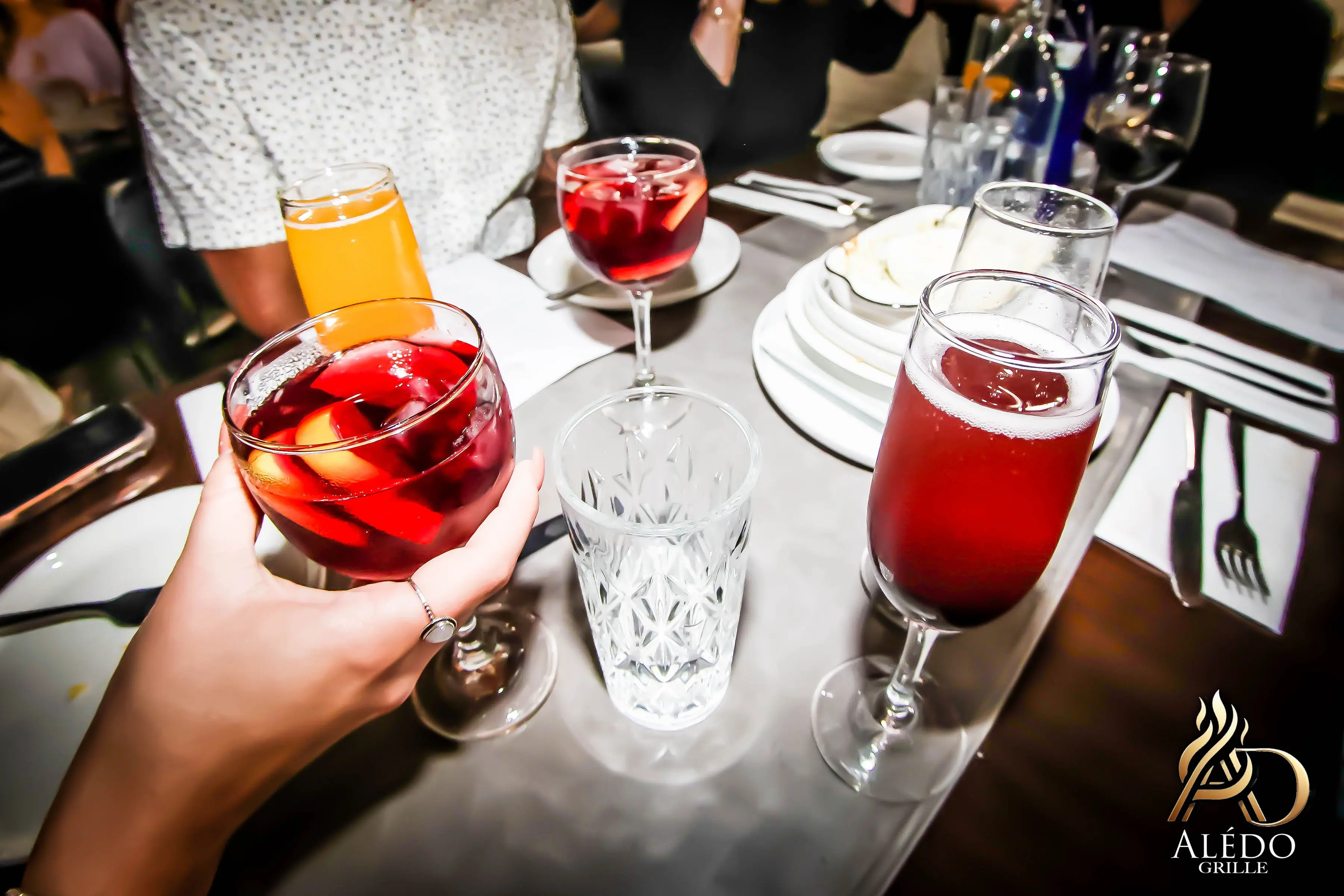 Hand holding a glass of red sangria with fruit pieces at a restaurant table with other drinks and place settings.