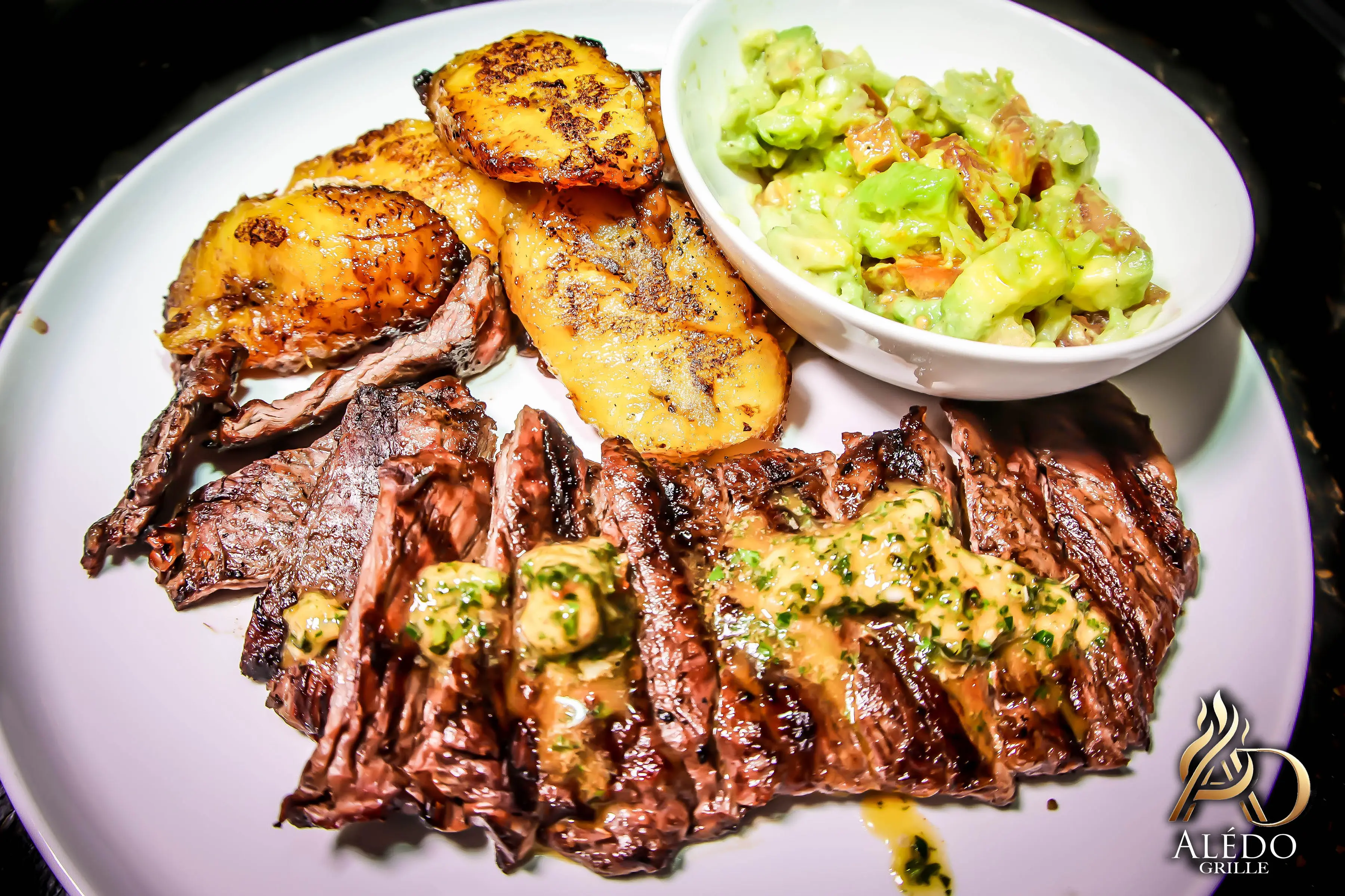 Grilled steak topped with herb butter, served with fried plantains and a bowl of avocado salsa on a white plate.