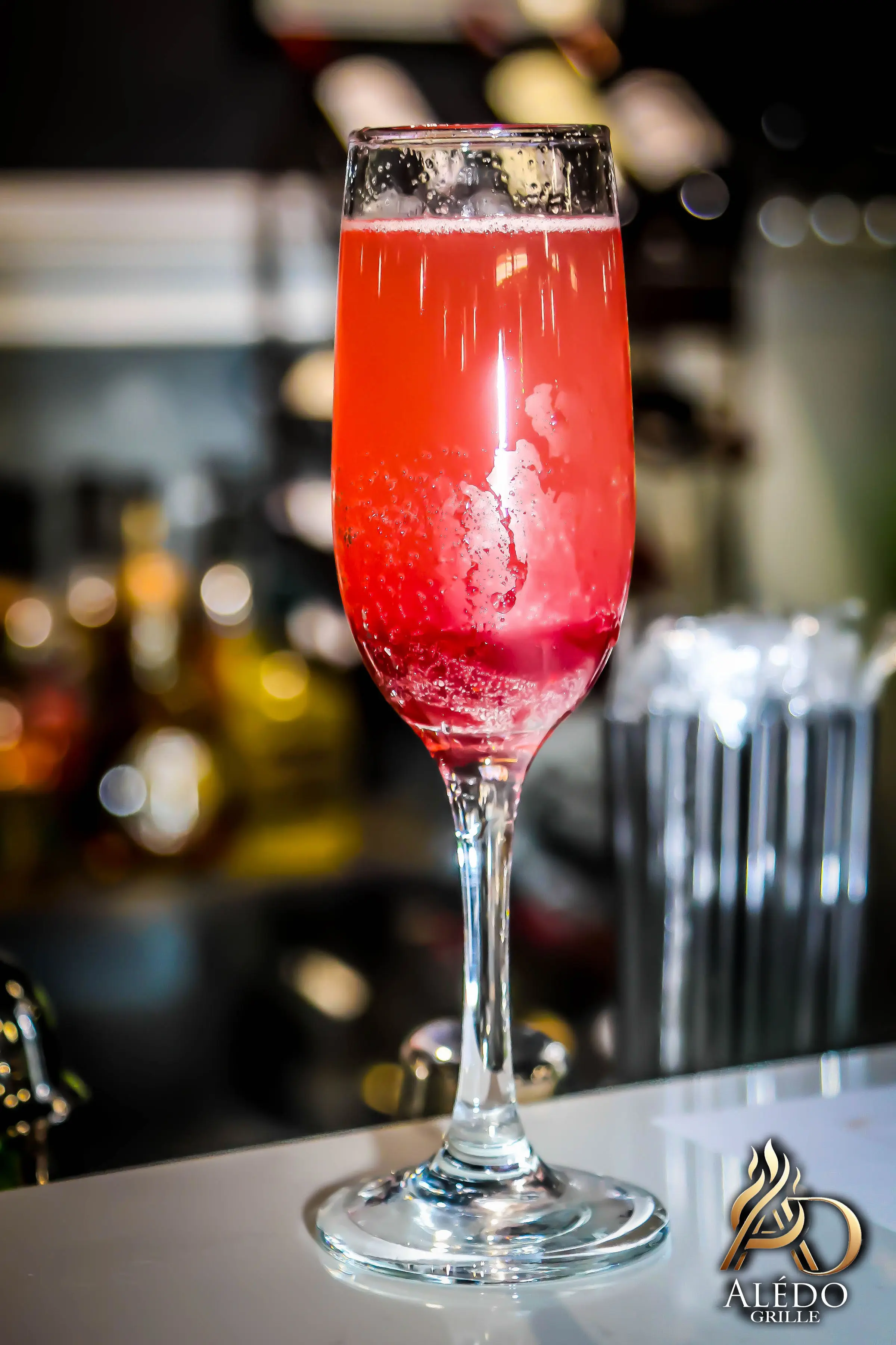Close-up of a red sparkling cocktail in a tall flute glass on a bar counter with a blurred background.