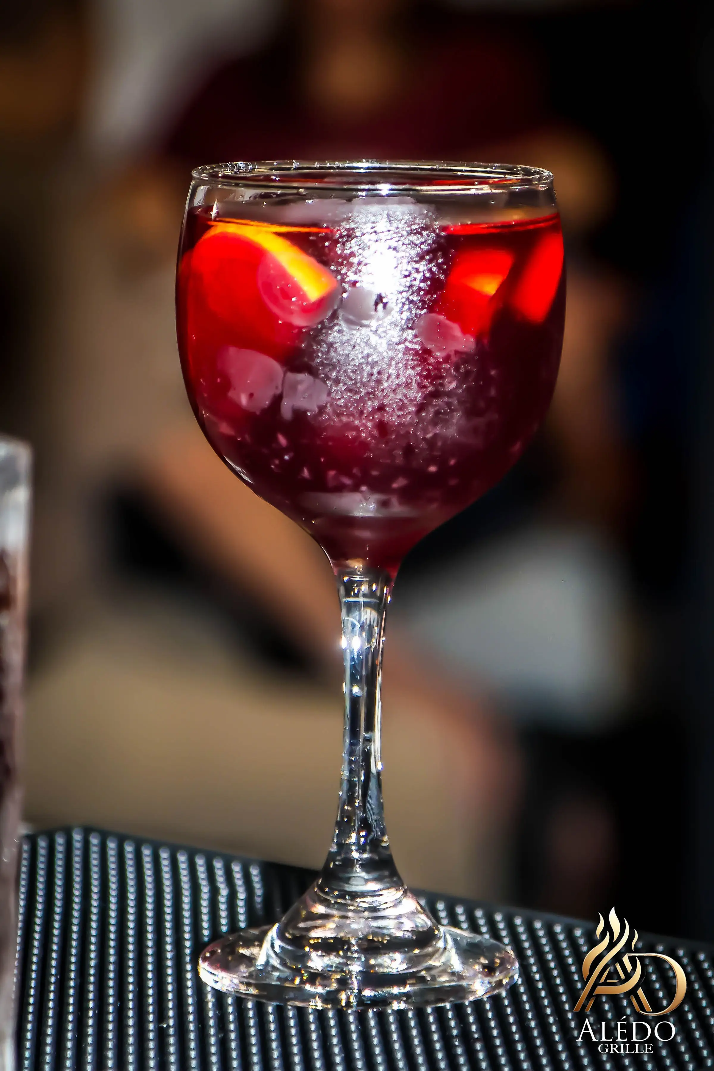 Chilled red cocktail with ice cubes and orange slices served in a stemmed glass on a textured surface.