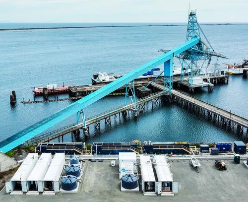 Industrial waterfront site with blue conveyor structure extending over piers into the water and various storage containers, tanks, and equipment onshore.