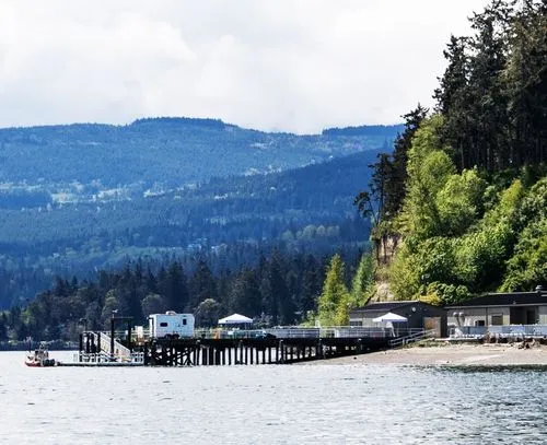 Wooden pier extending into calm water with a white trailer and tents, backed by dense green trees and forested hills under a cloudy sky.