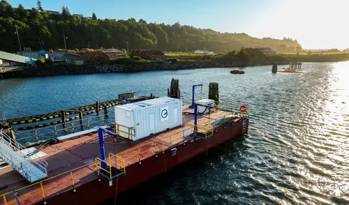 Large industrial barge docked on calm water near a green hillside and stacks of cut logs.