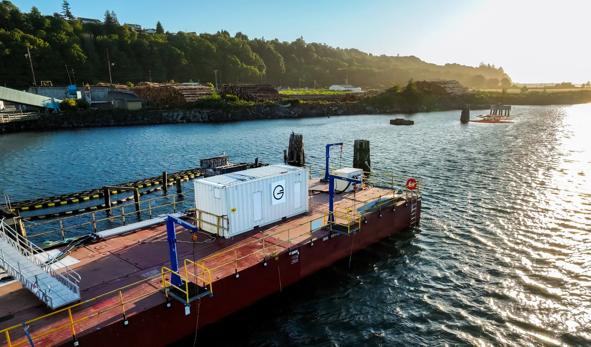 Large industrial barge docked on calm water near a green hillside and stacks of cut logs.