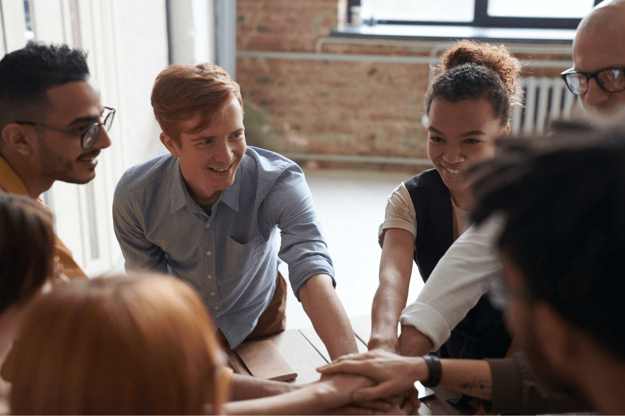 A group of coworkers are smiling and putting their hands together in a circle. The wall behind them is brick, and there is a white heater on the wall under a window.