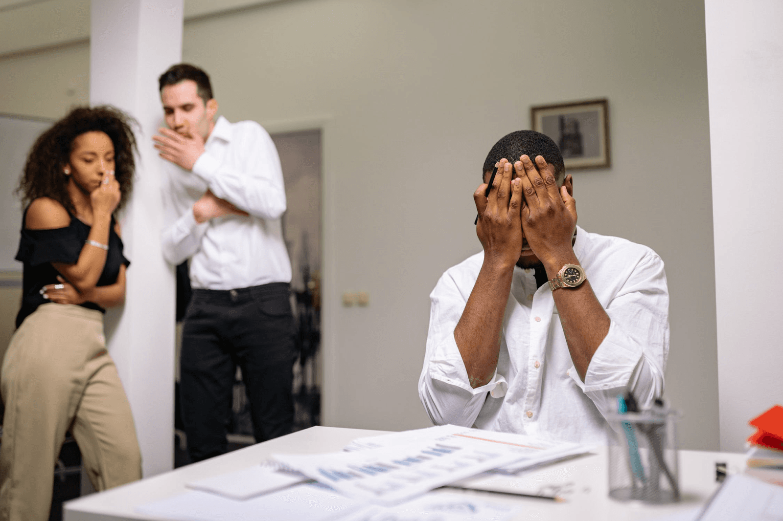 A worker is sitting at their desk and has their head in their hands. There are two people standing in the background, and they are talking to each other. There are papers and a jar of pens on the desk.