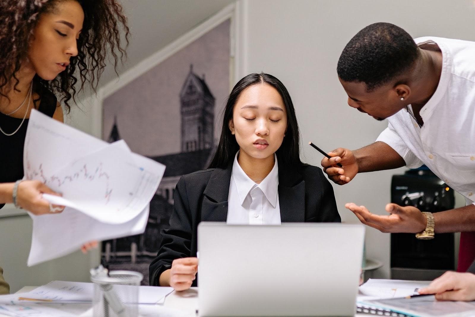 A person is sitting at their computer is looking frustrated as one person is talking to her and another person is showing her graphs and charts. The frustrated person is wearing a white button-up and a black blazer.