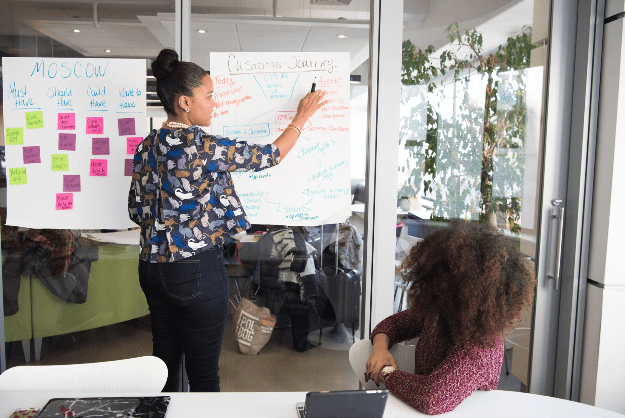 Two people are in a meeting, one is standing up and teaching the other about the customer journey. The person standing has on a multi-colored, patterned top and jeans. The person sitting down has a purple sweater on.