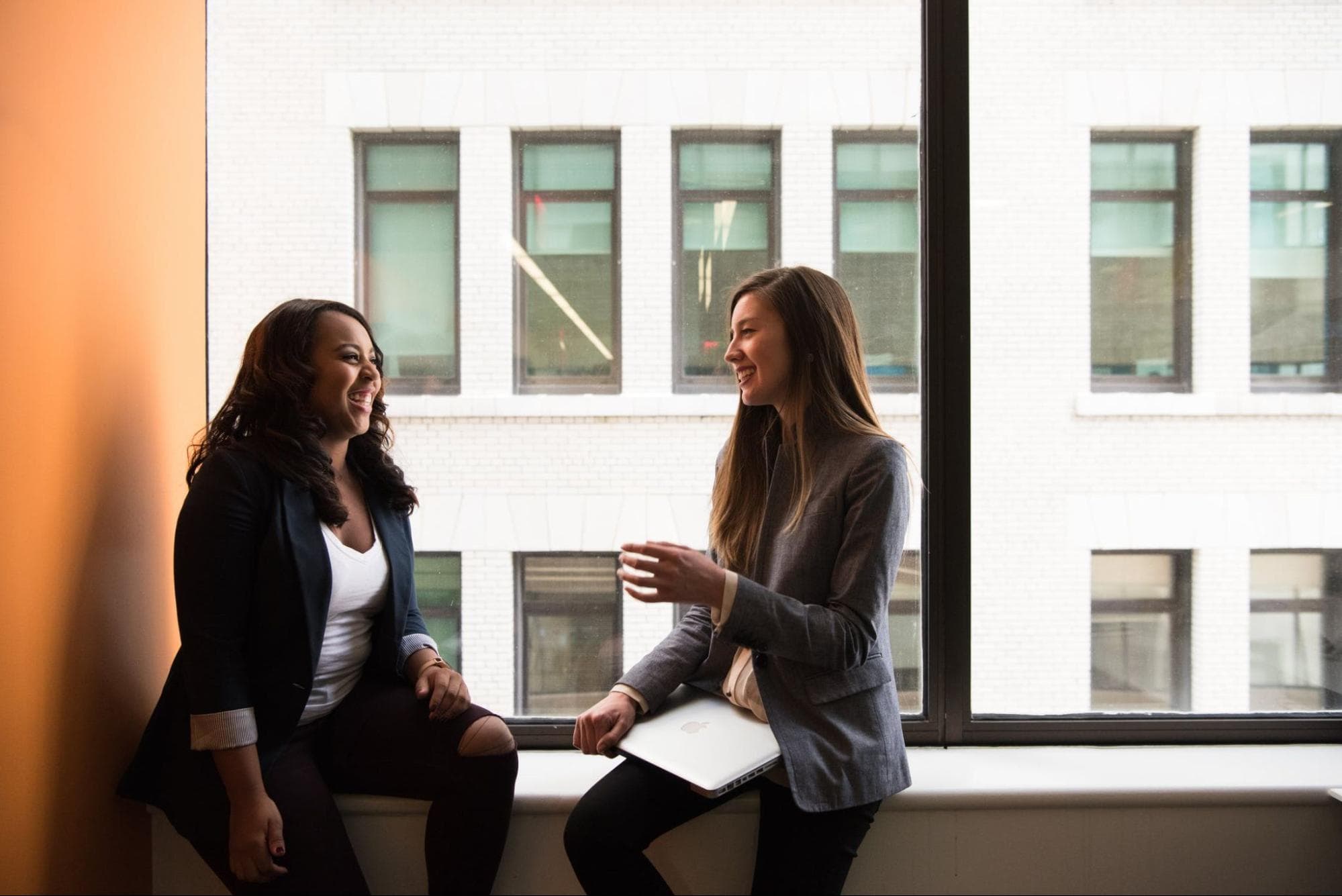 Two people are sitting on a window sill talking to each other. One person has a laptop in their lap. They are both wearing dark-colored blazers and dark pants