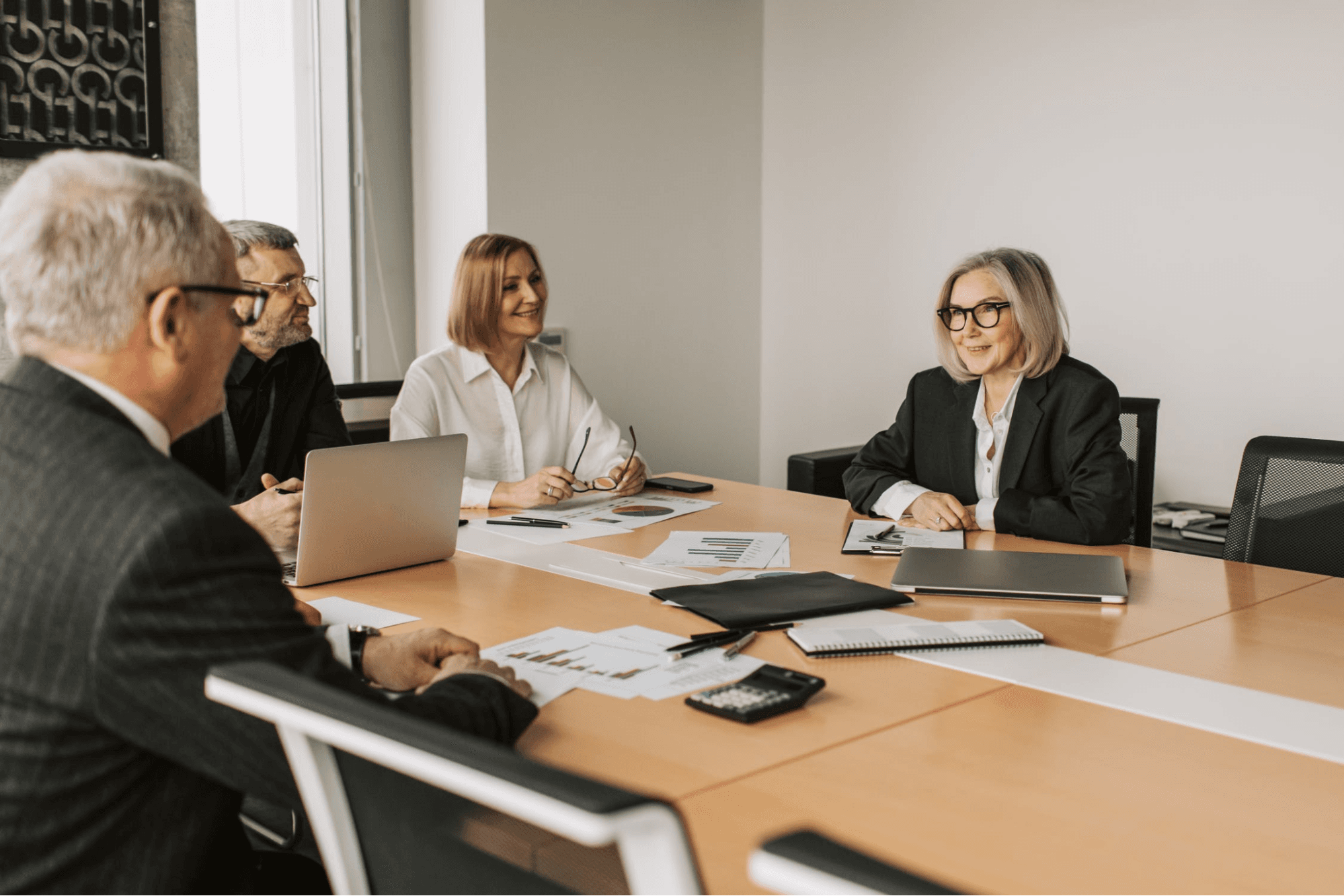 Four people are sitting around a table. The table has computers and papers on it. The people are all wearing black and white clothing. Three people are wearing glasses.
