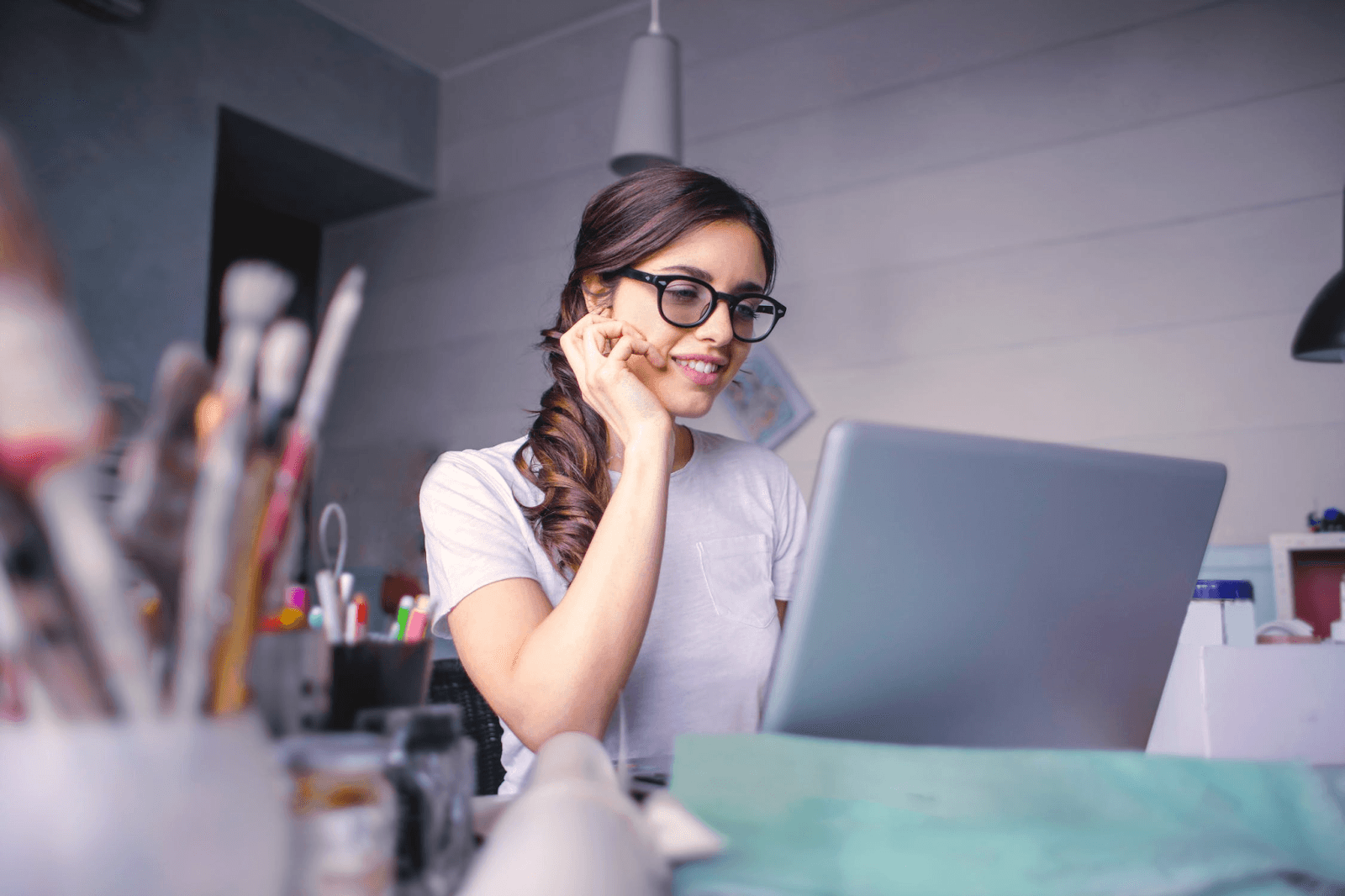 A person sitting at their desk looking at their silver laptop. They are wearing glasses and have their hair in a ponytail.
