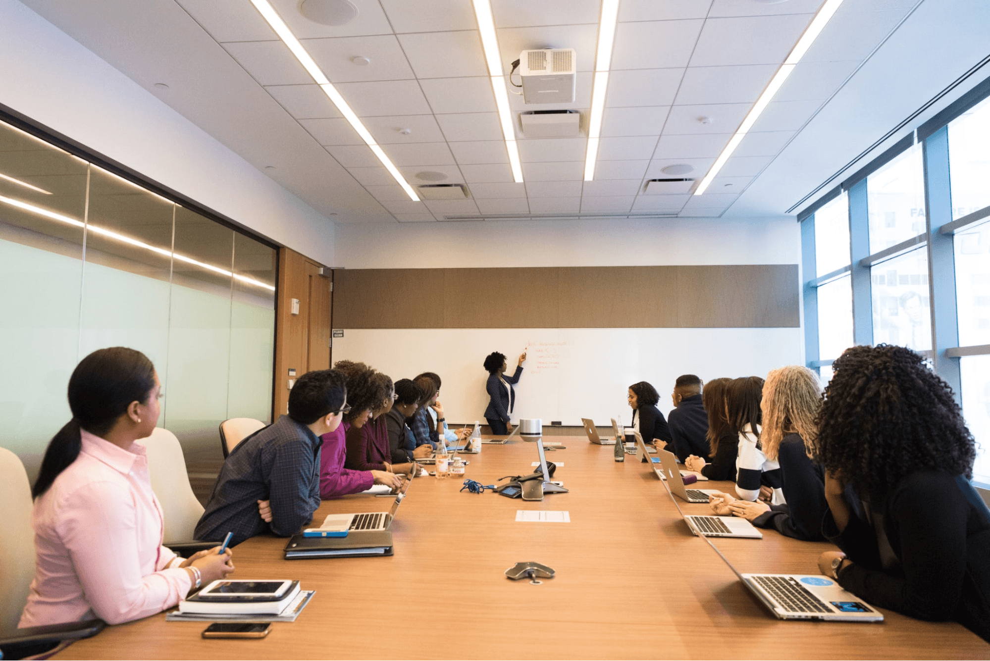 A large meeting is being held in a room with a long wooden table with a large whiteboard. Various people have their notebooks, laptops and phones on the table.