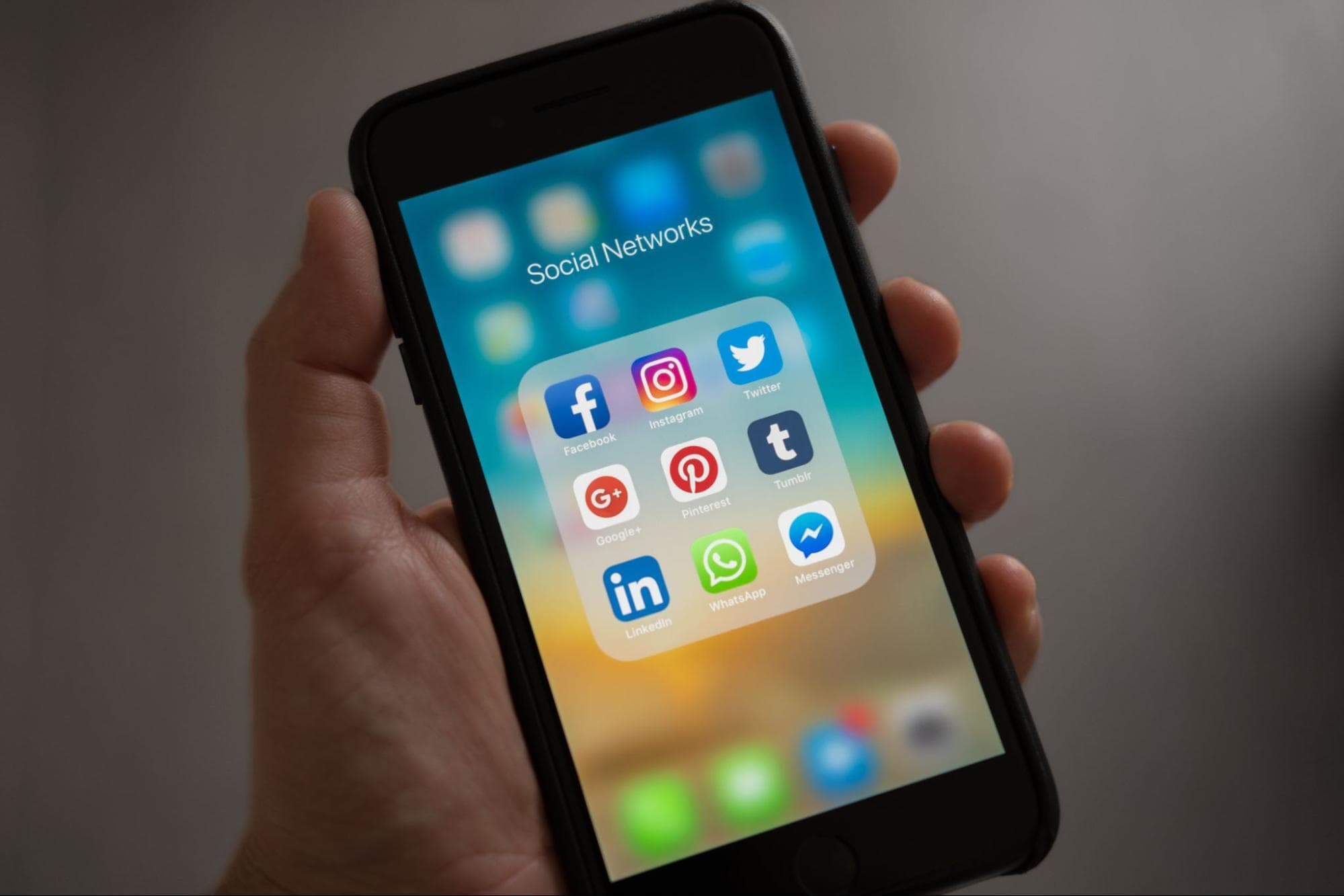A person is holding their phone. They have a “social networks” folder with various apps in it, including Facebook, Instagram, Twitter, Pinterest, LinkedIn, Facebook Messenger, WhatsApp, Google+ and Tumblr.