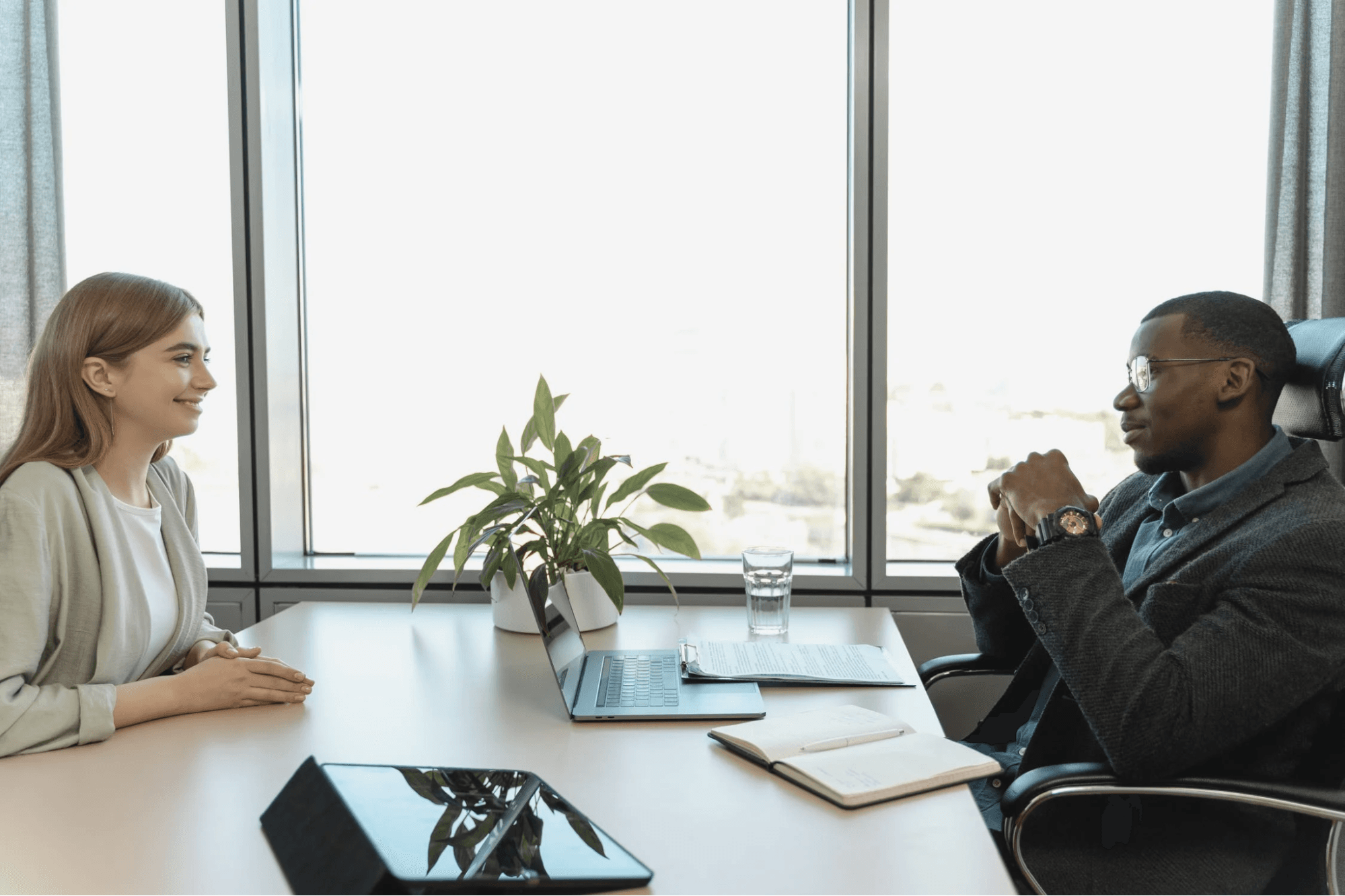 This is a one-on-one with two employees. The table has a plant, a laptop, a notebook and a water glass on it. One employee is wearing a dark jacket, a blue button down, glasses and a watch. The other employee is wearing a white shirt with a tan cardigan.