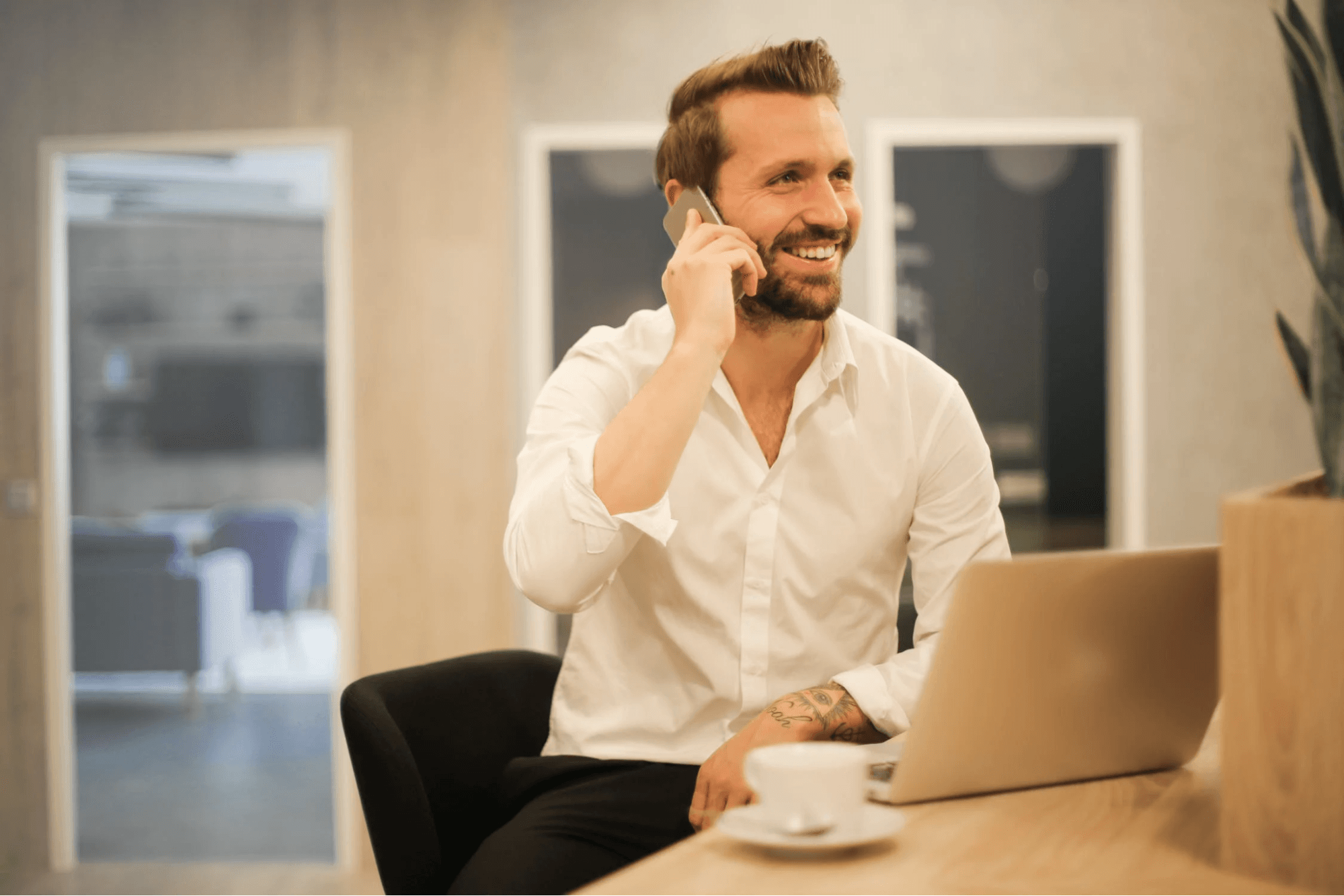 A person is talking on the phone. They are sitting at a table with a laptop and drinking a cup of tea. They are wearing a white shirt and black pants