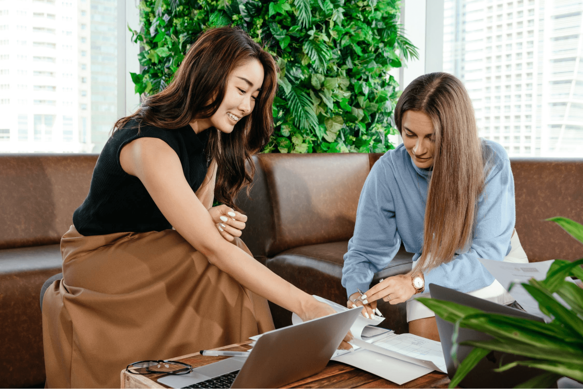 Two people are sitting on a couch looking over some papers. They are sitting at a coffee table with a laptop. One person is wearing a black shirt and a tan skirt, and the other is wearing a blue sweater and a white skirt. There are various plants behind them.
