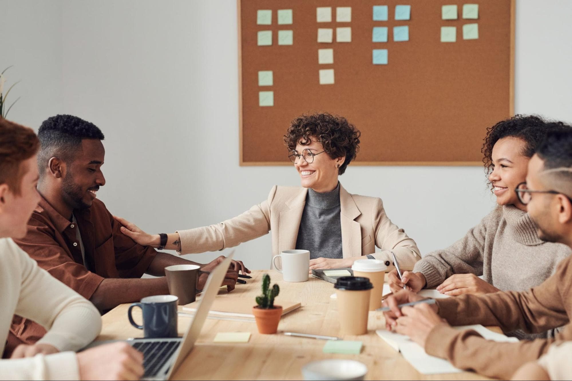 A group of people sits around a table talking and smiling. They are all wearing neutral color clothes, and there is a corkboard behind the table with notes pinned to it.