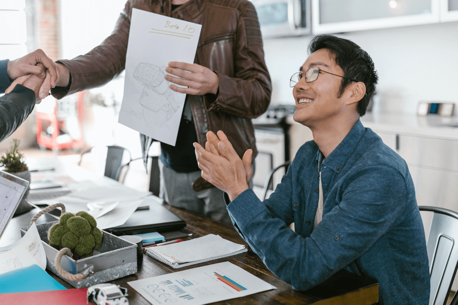 A person wearing a blue jacket is at the office and clapping their hands. They are sitting in a chair. The person standing next to them is wearing a brown jacket and shaking someone’s hand.