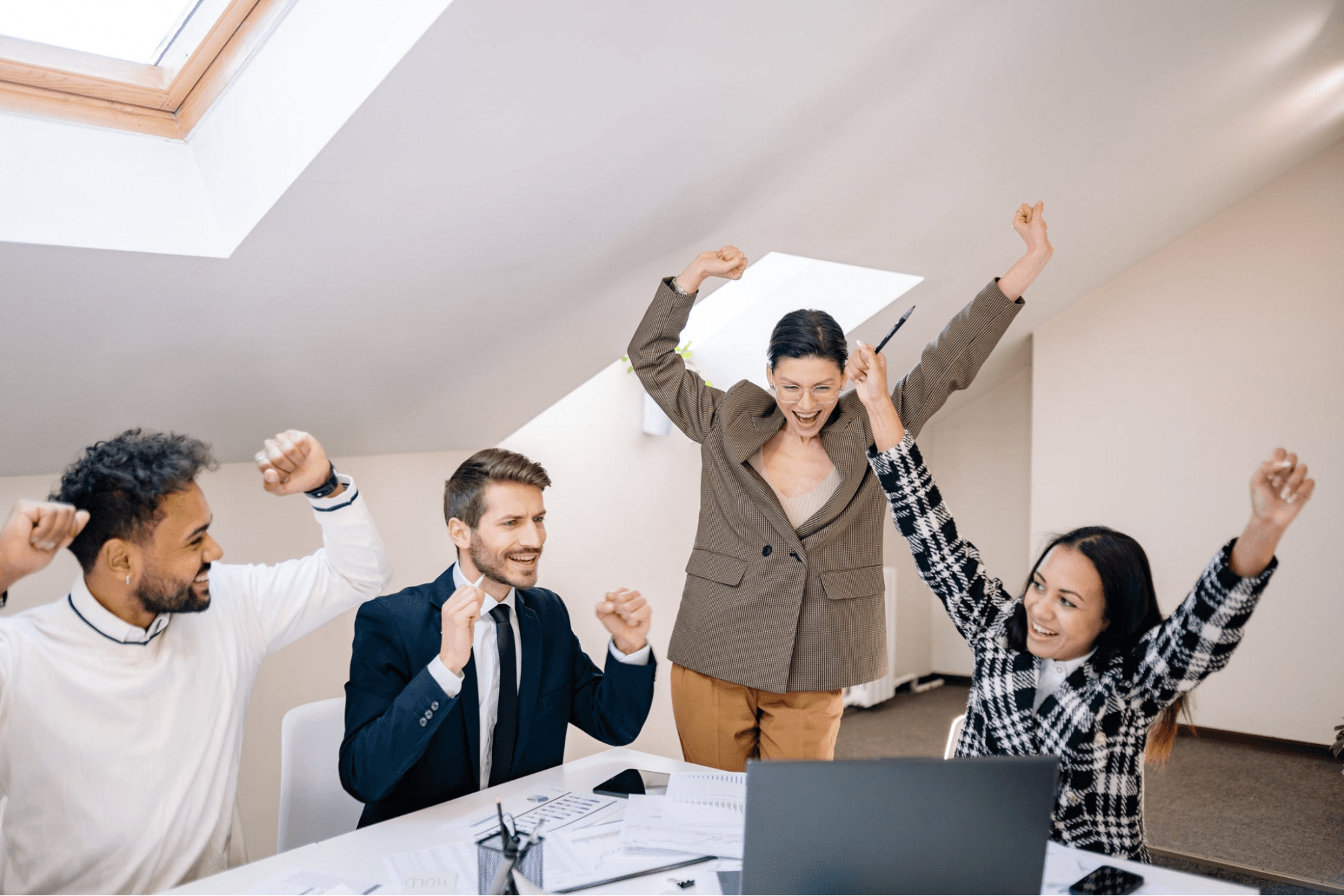 Four people are celebrating with their arms in the air and smiles on their faces while there are papers and a laptop are sitting on the table.