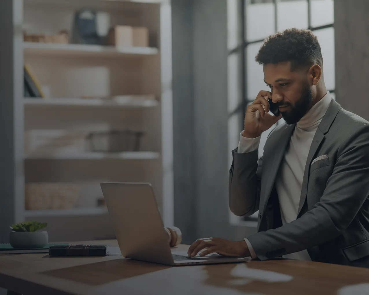A black man sits at his desk in front of his laptop while talking on his smartphone