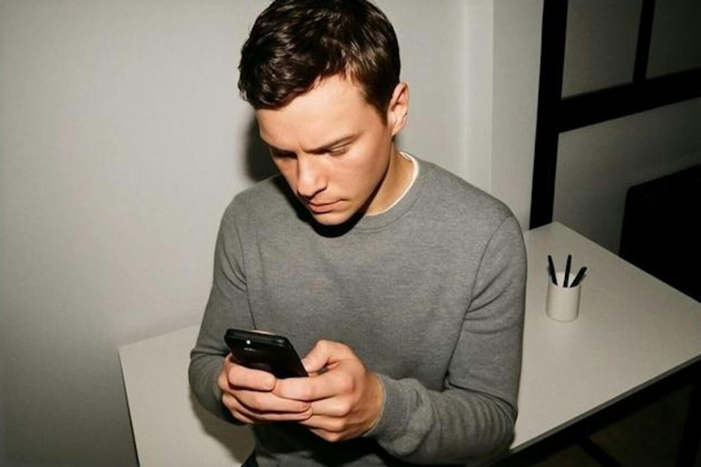 Man in a grey sweater scrolling on his phone at a white desk.