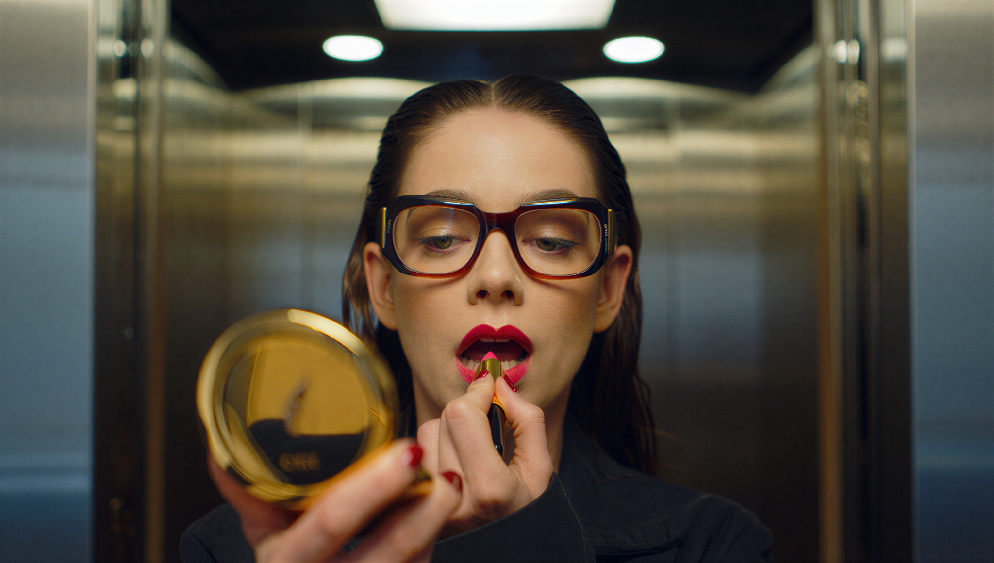 Woman in oversized glasses applying red lipstick with a gold compact mirror in an elevator