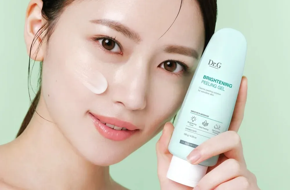 Woman holding a brightening peeling gel tube beside her face with a dab of gel on her cheek