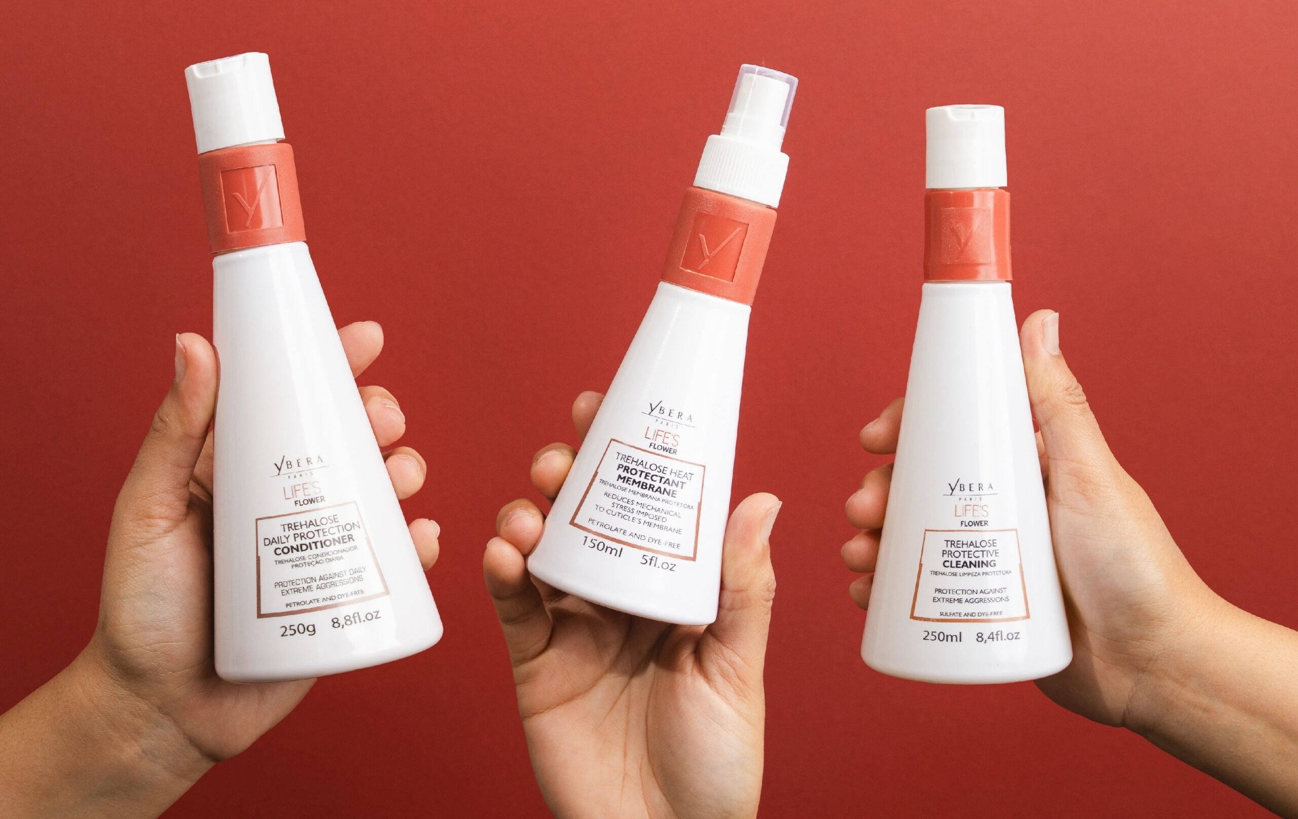 Three hands holding white hair care bottles with coral caps against a red background