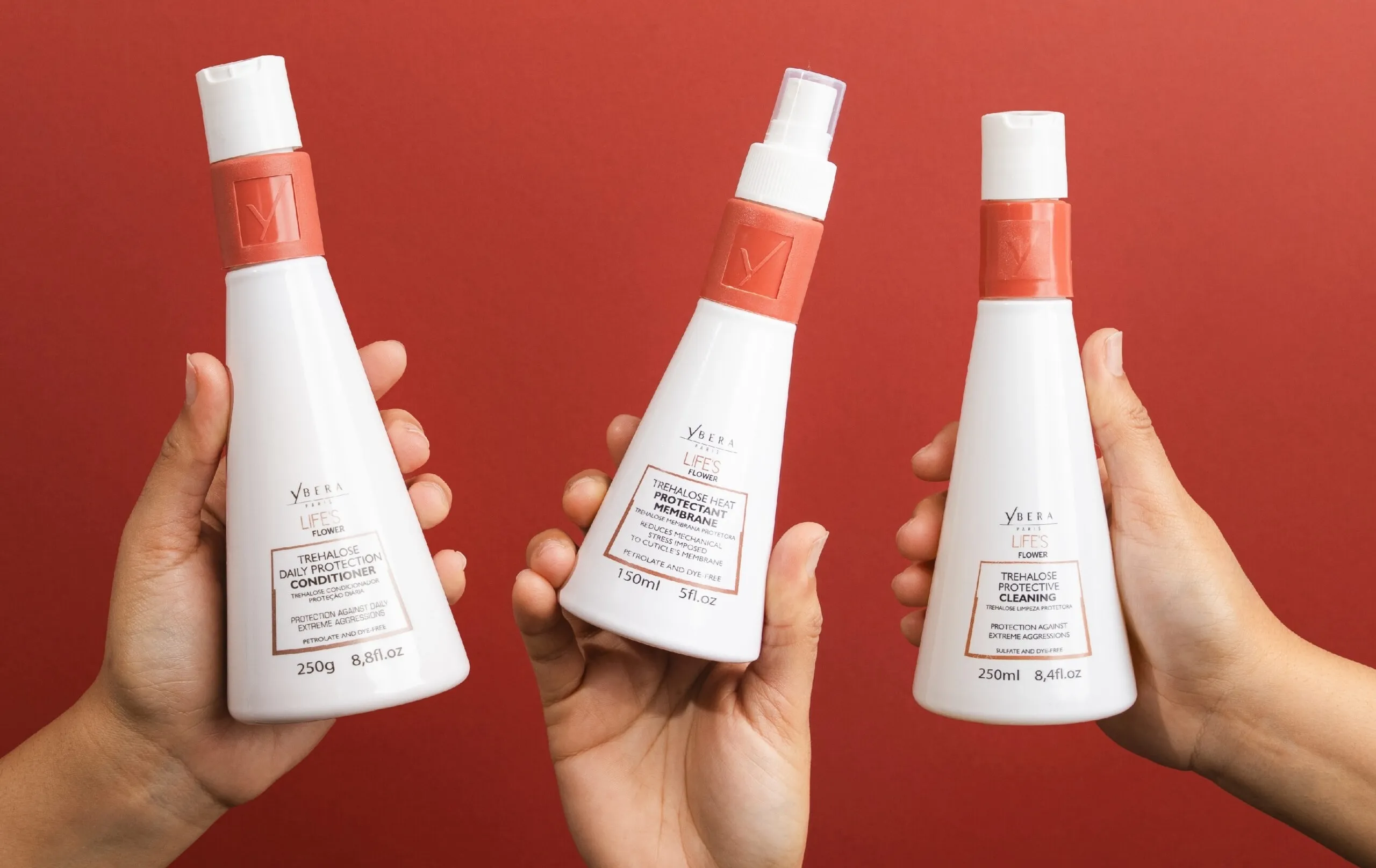 Three hands holding white hair care bottles with coral caps against a red background