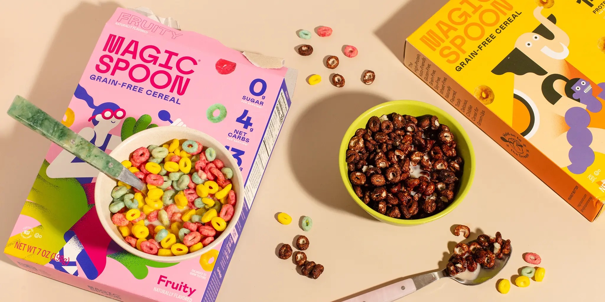 Bowls of fruity and chocolate grain-free cereal beside colorful cereal boxes on a cream tabletop