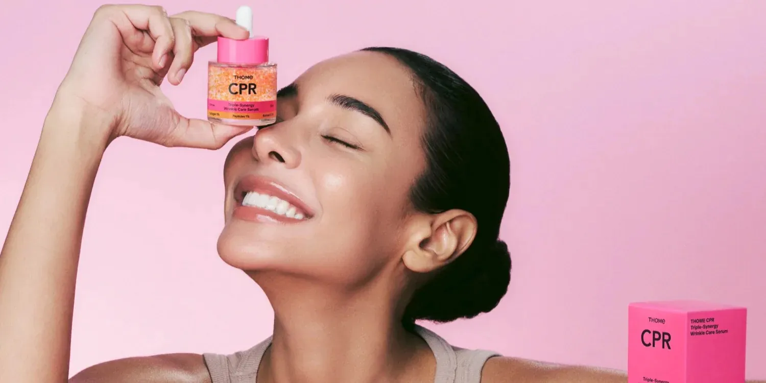 Smiling woman holding a wrinkle care serum dropper bottle to her eye against a pink background