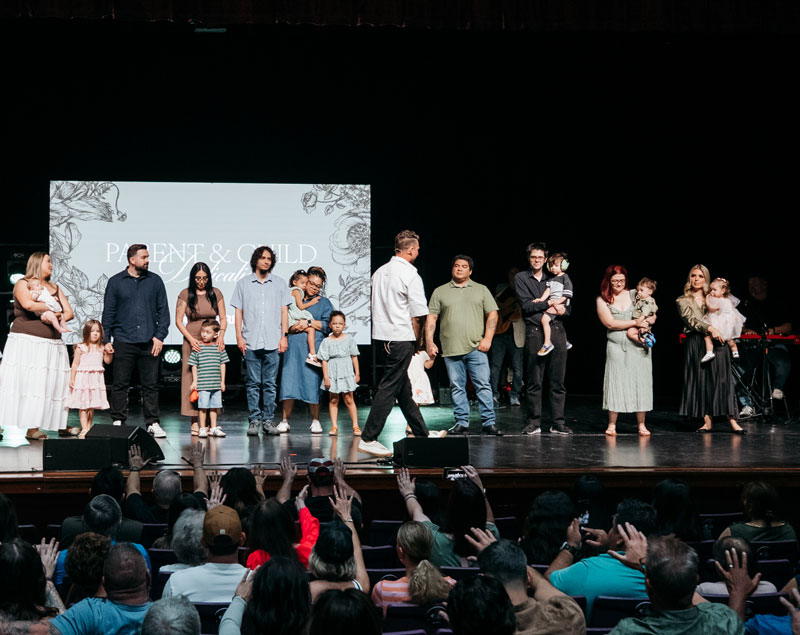 Parents and children stand on stage at a Parent & Child event with an audience raising hands.