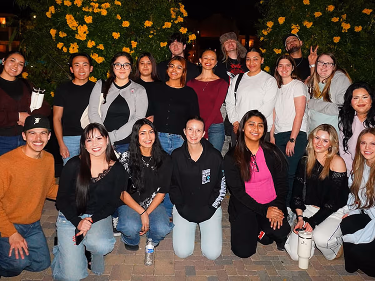 Group of diverse young adults posing outdoors at night with yellow flowers in the background.