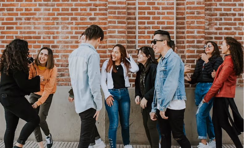 Group of eight diverse young adults chatting and laughing by a brick wall outdoors.