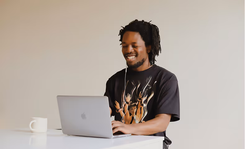 Smiling man with dreadlocks using a laptop at a white table with a white mug nearby.