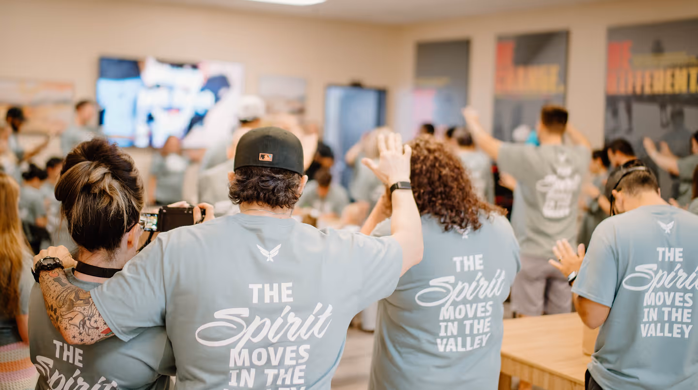 Group wearing matching blue shirts with 'The Spirit Moves in the Valley' raising hands in a room.