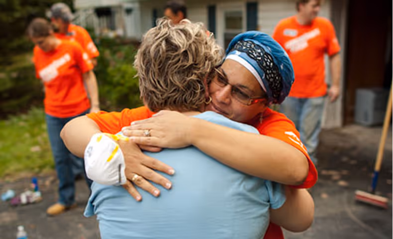 Two women warmly hugging outdoors while people in orange shirts work in the background.