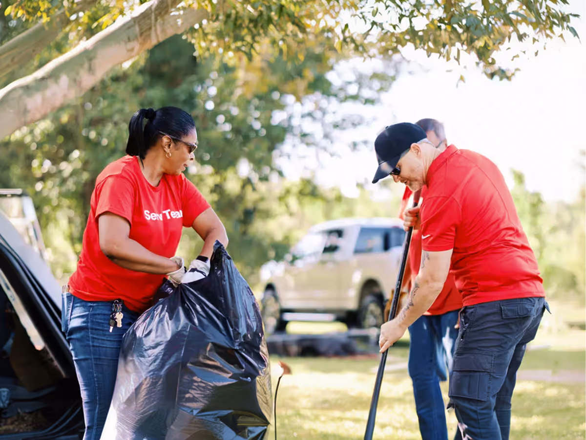 Volunteers wearing red shirts clean up outdoors, one holding trash bag and another with rake.