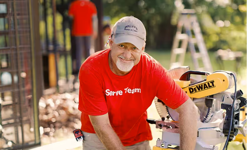 Smiling man in red Serve Team shirt and cap using a DeWalt miter saw outdoors.