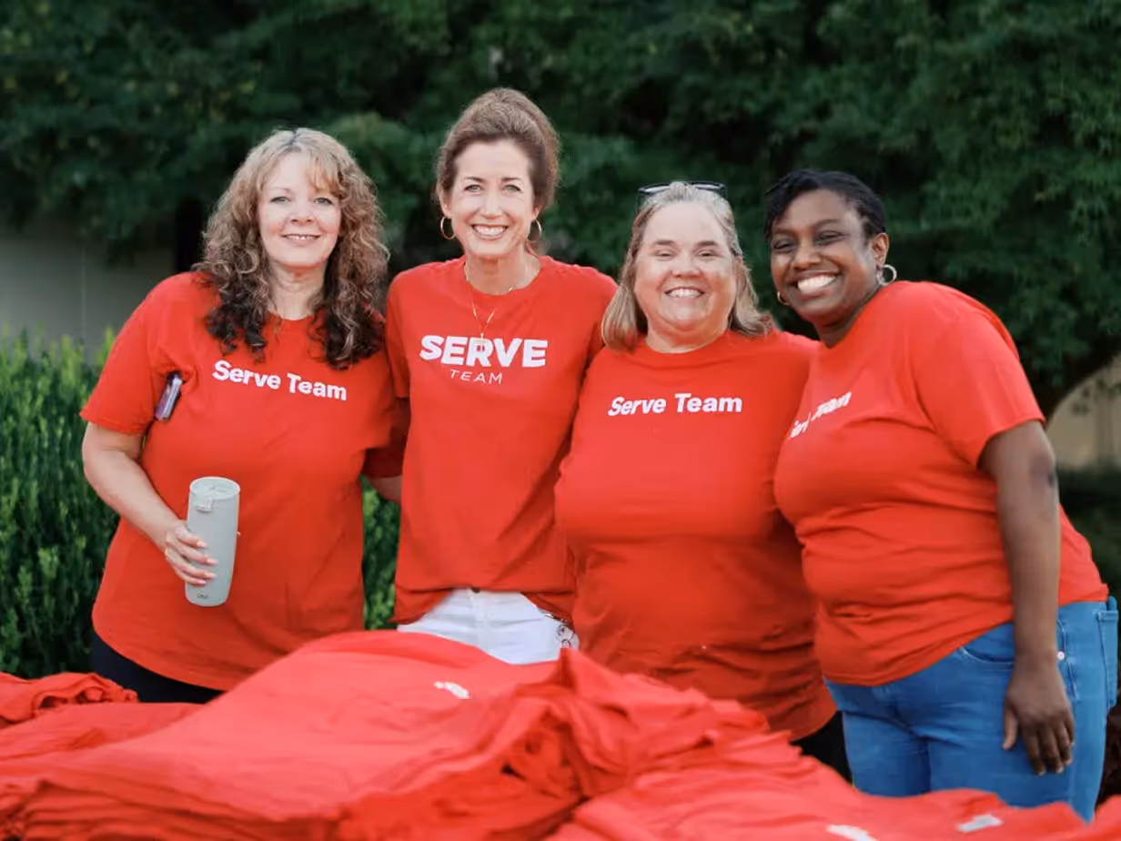 Four smiling women wearing red Serve Team shirts, standing outdoors with trees in the background.