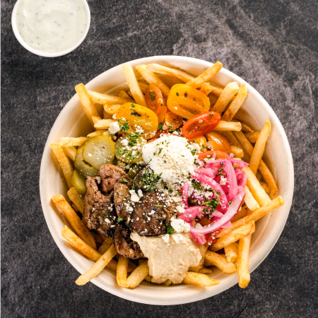 Bowl of seasoned beef slices on french fries topped with cherry tomatoes, pickles, pickled red onions, feta cheese, hummus, and a side of creamy sauce.