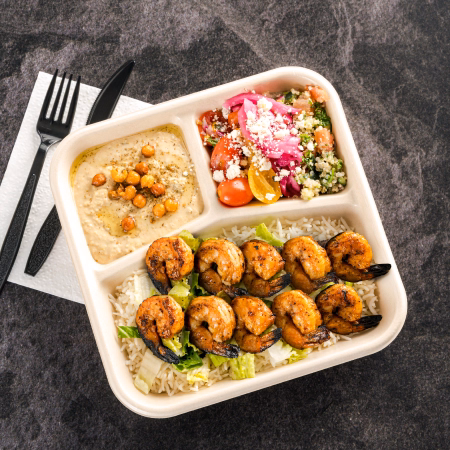 Three-compartment meal tray with grilled shrimp over rice and lettuce, creamy hummus topped with roasted chickpeas, and a quinoa salad with tomatoes, pickled onions, and feta cheese.
