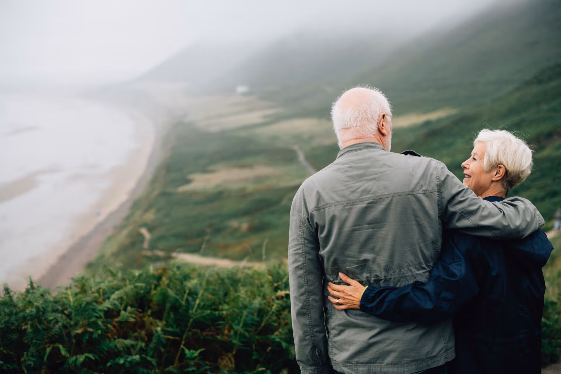 An older couple with their arms around one another looking over a foggy bay.