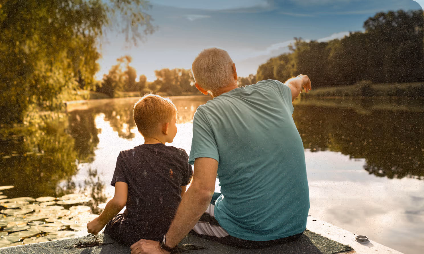 a child and his grandpa sitting next to a pond at sunset looking into the distance