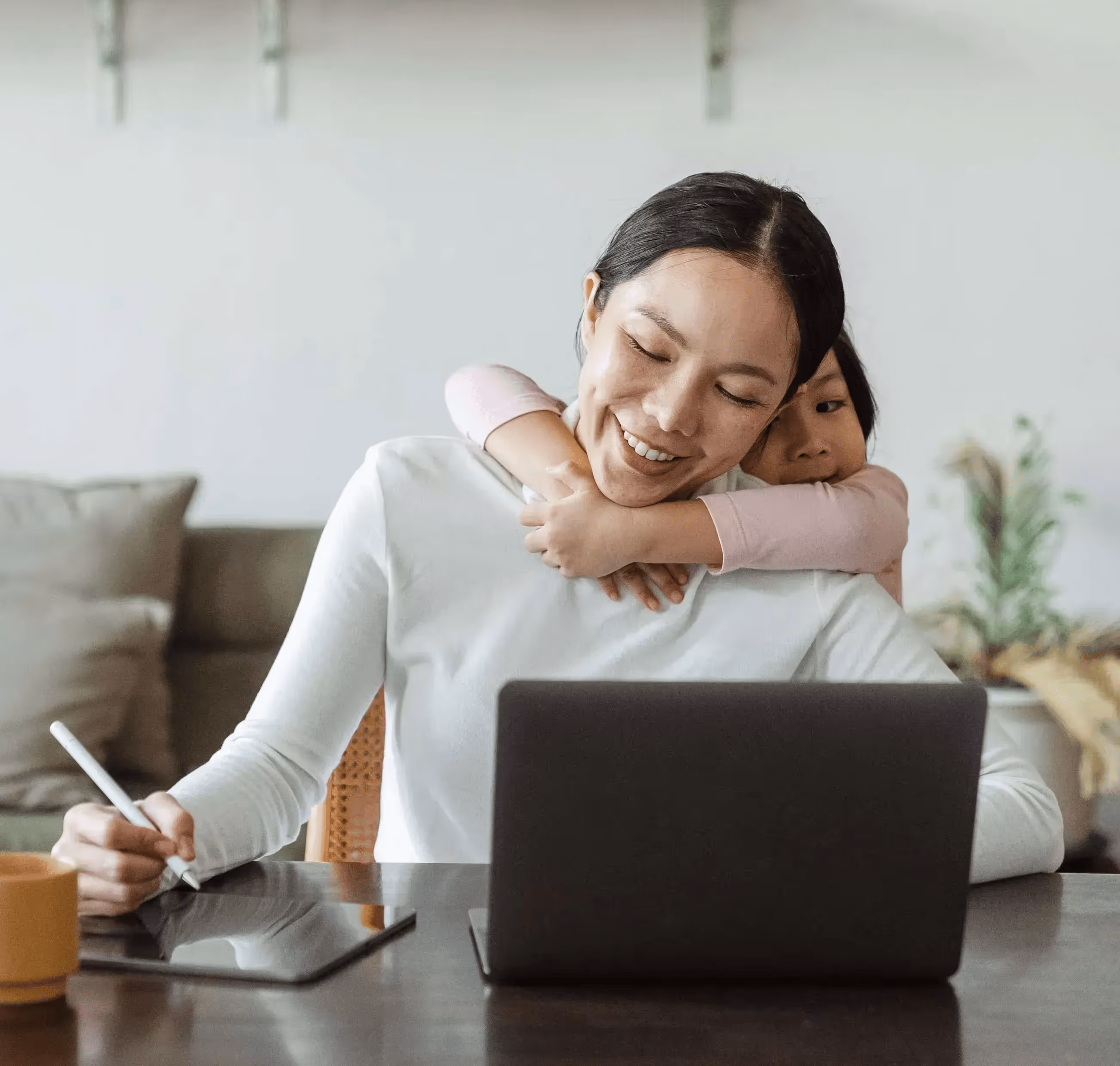 A mother on her laptop with a notepad writing things down with her child hugging her from behind